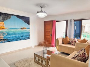 Un salón con un cuadro grande de mar, una araña, un sofá con cojines estampados y una mesa de centro de cristal.