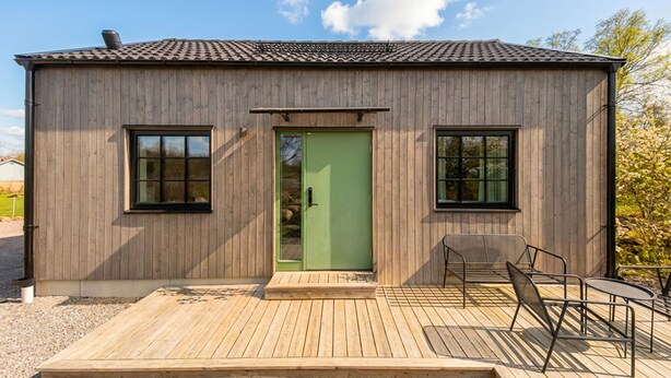 Ein kleines Holzgebäude mit einer grünen Tür, zwei Fenstern und einer Terrasse mit Metallstühlen.