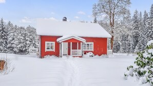 En röd stuga med en vit veranda, omgiven av snötäckta träd.