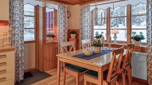 Ett mysigt matrum med träpanel och ett bord dukat för fyra personer, omgivet av stolar. På fönsterbrädan står krukväxter och ovanför bordet hänger en kristallkrona.