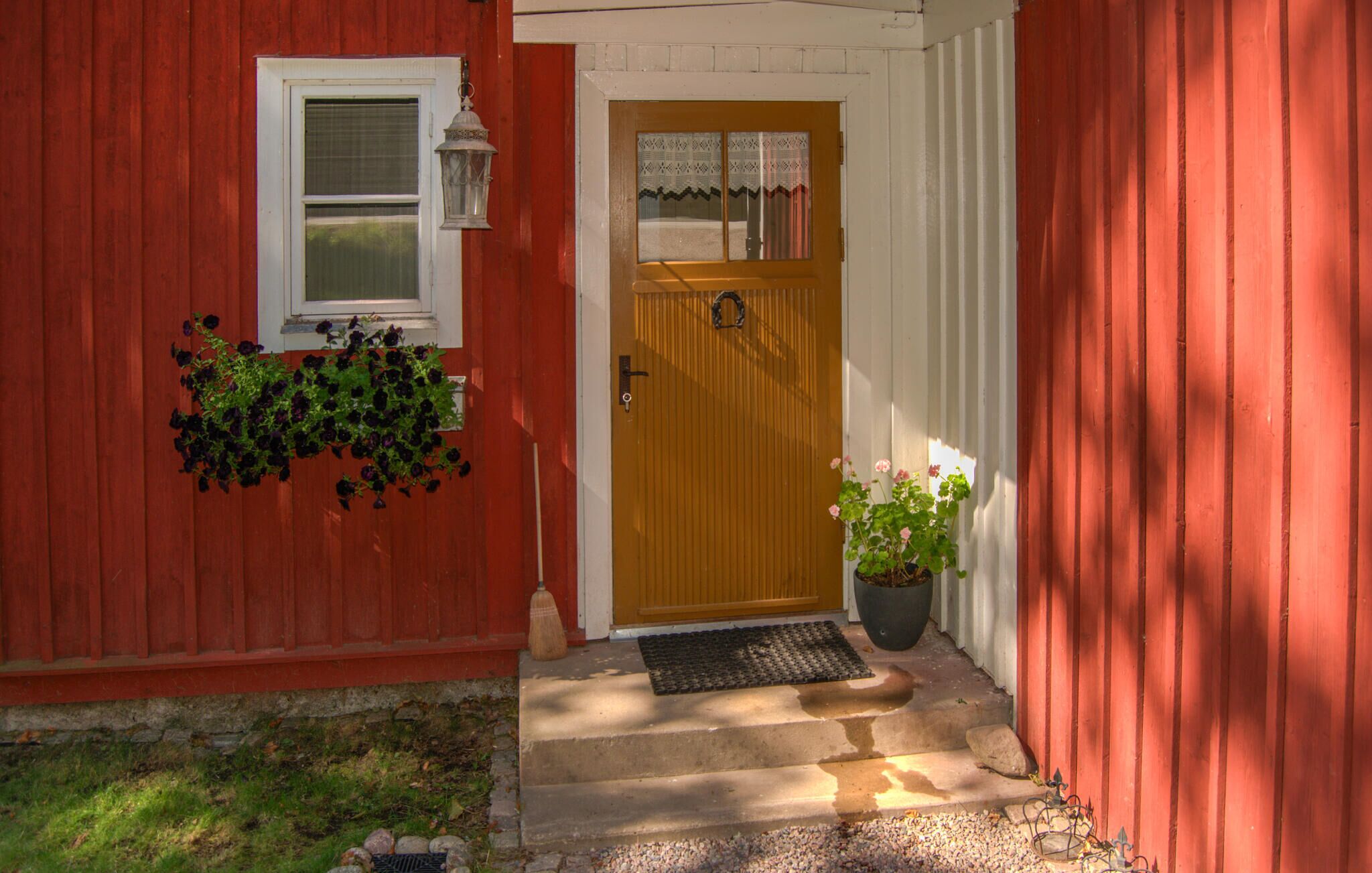 A red wooden house with a white door, a window with a white frame, and a potted plant with pink flowers.
