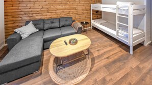 Une pièce comprenant un canapé d’angle gris, une table basse ronde en bois sur laquelle est posé un plateau avec des plantes, un lit superposé blanc et un mur en bois.
