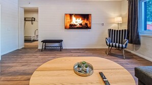 Un salon moderne avec une table basse ronde en bois, un banc, un fauteuil à rayures et un téléviseur à écran plat fixé au mur, qui affiche une image de cheminée.