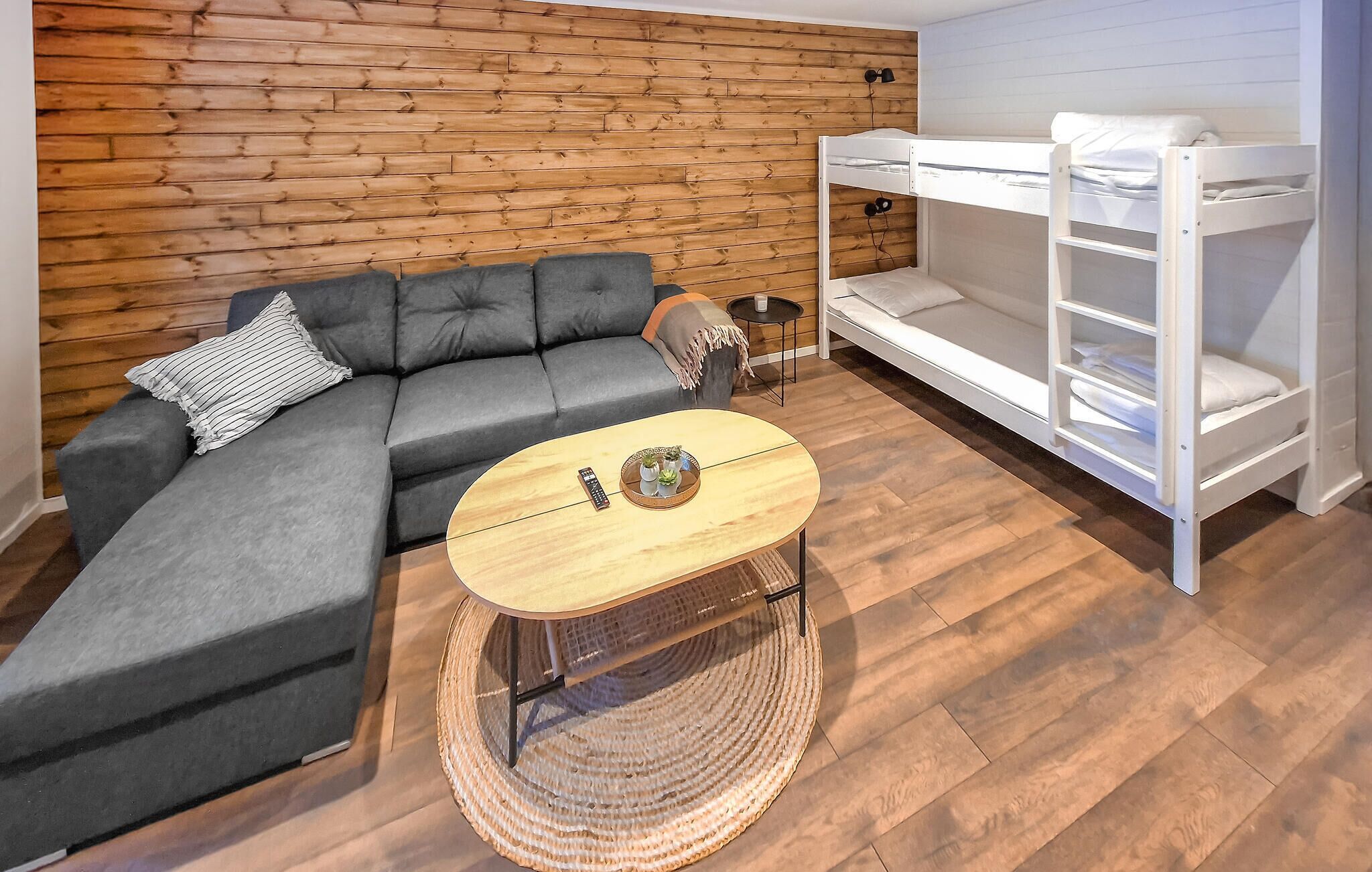 A room with a grey sectional sofa, a round wooden coffee table with a tray of plants, a white bunk bed, and a wooden wall.
