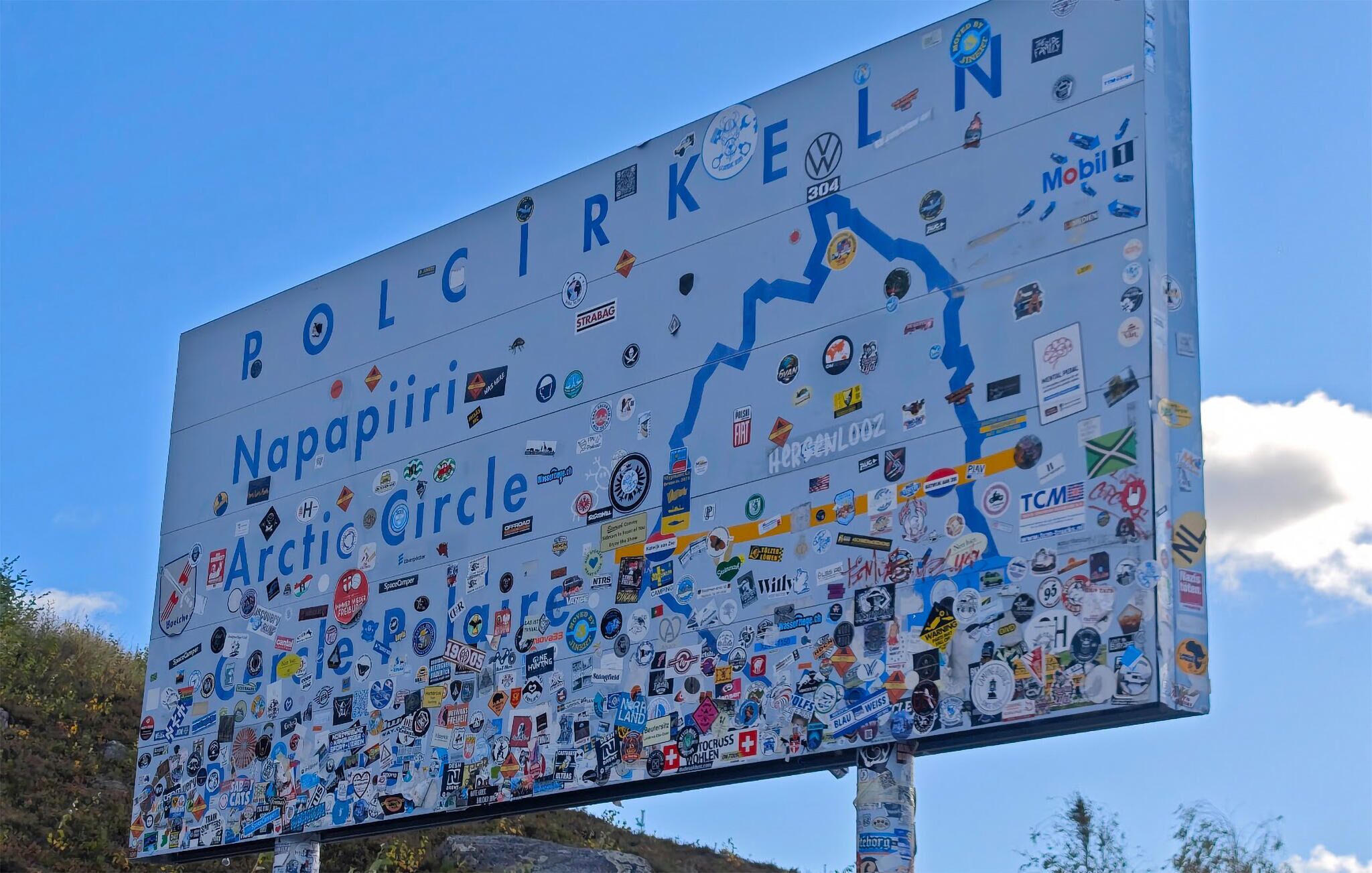 A large sign with the text "Nappalpiiri Arctic Circle" surrounded by numerous stickers.