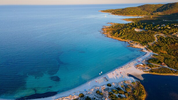 Luchtbeeld van een kustgebied met een strand, helderblauw water en omliggende begroeiing.