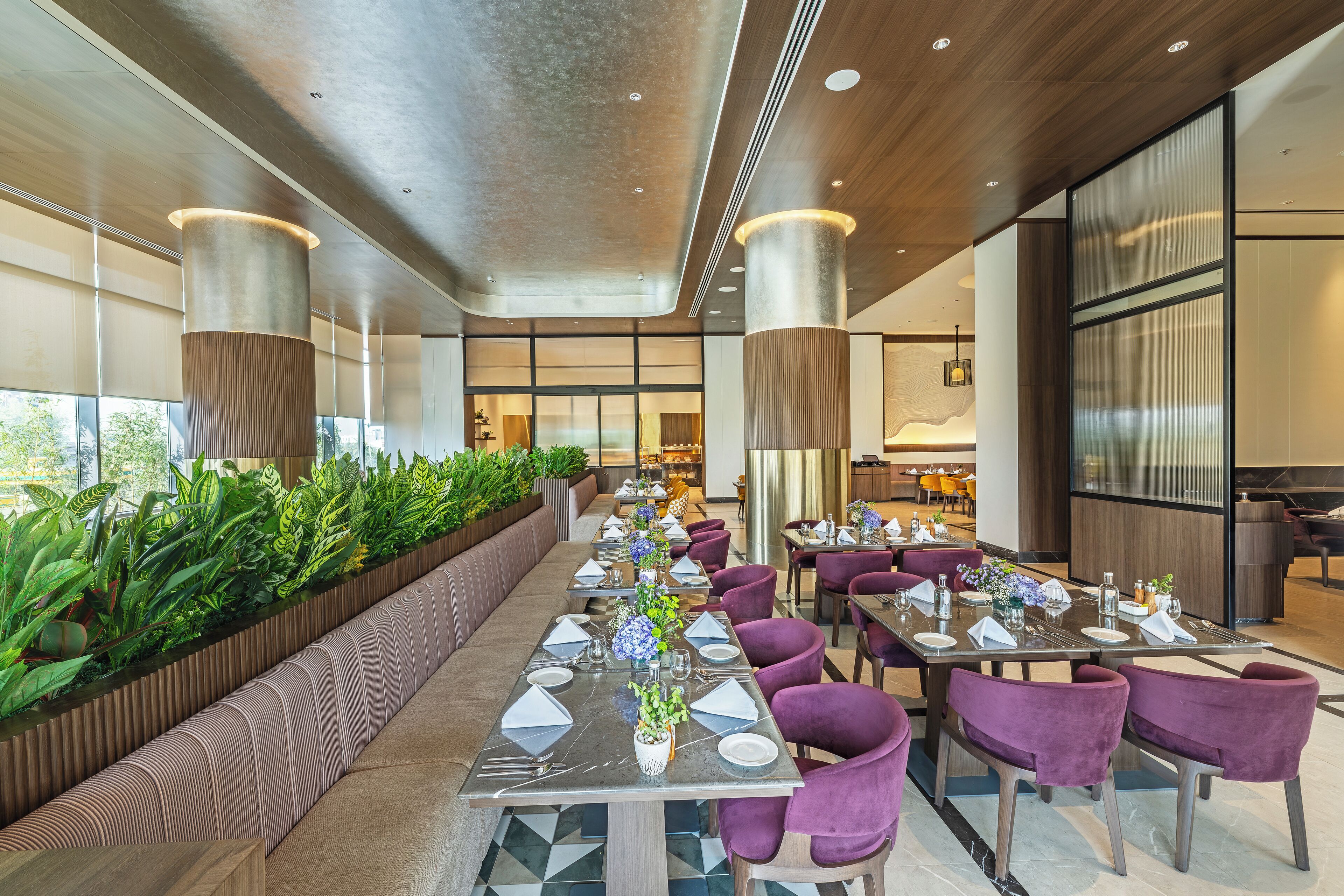 A modern dining area with long tables, purple chairs, and a green plant wall.