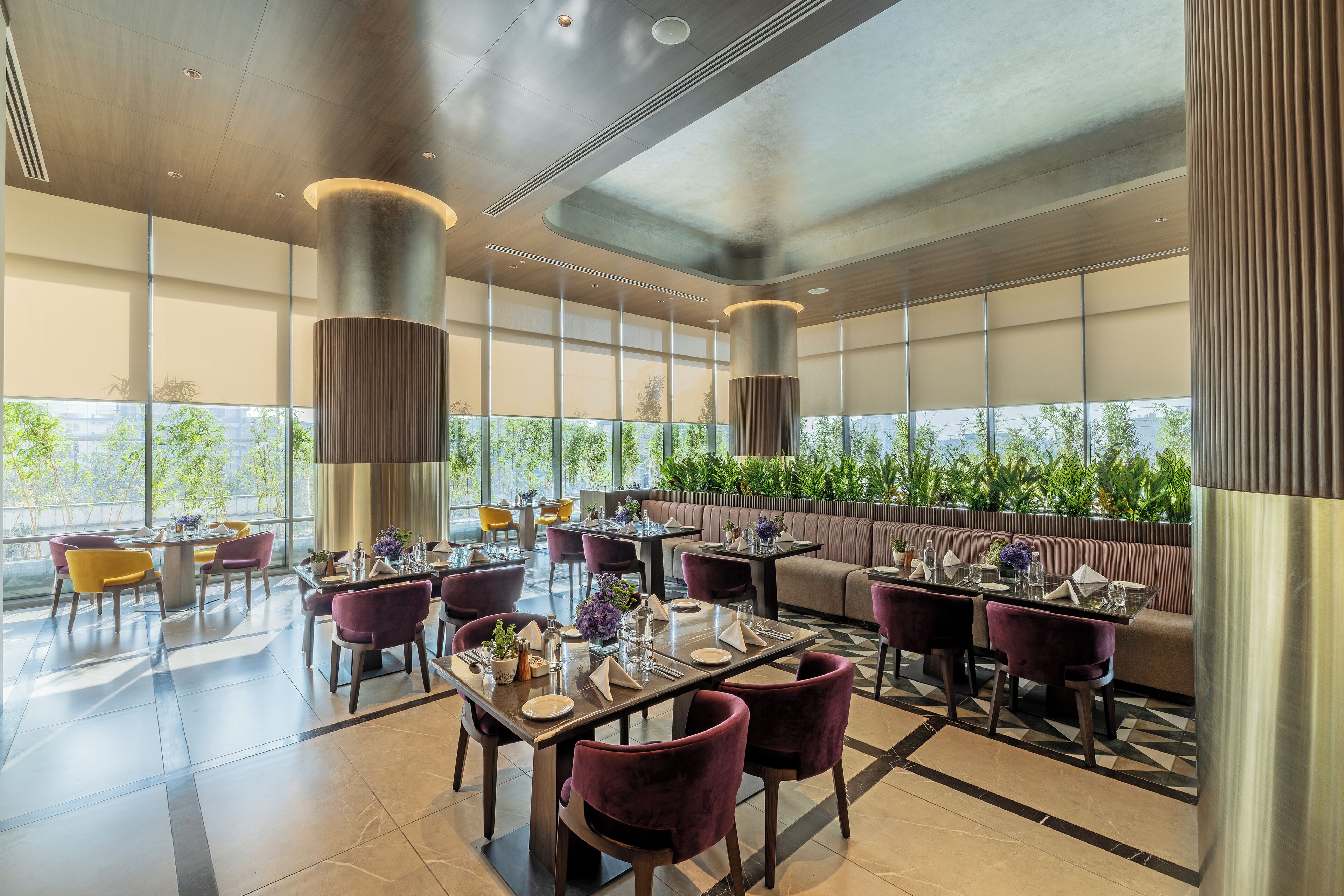 A modern dining area with tables and chairs, large windows, and a view of greenery outside.