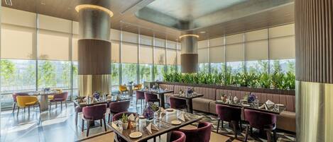 A modern dining area with tables and chairs, large windows, and a view of greenery outside.