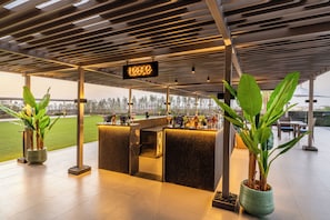 A modern outdoor bar area with a sign, potted plants, and a view of a green field. - Vivanta Vrindavan (Vrindavan)