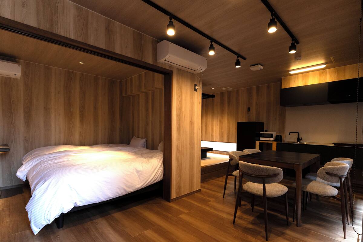 A modern room with a bed, a dining table with chairs, a kitchen area, and built-in wooden walls.