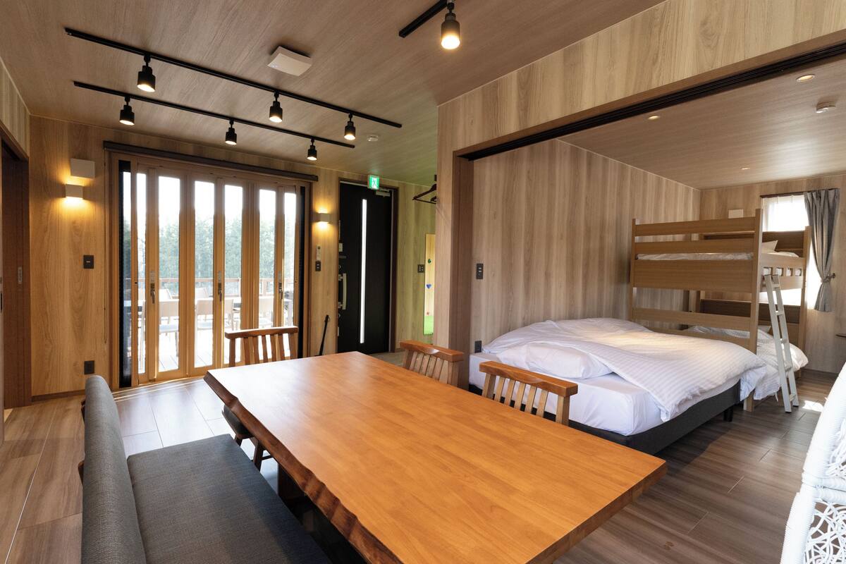 A modern hotel room with a wooden floor, a large wooden table, a bunk bed, and a sliding glass door leading to a balcony.