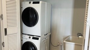 Uma lavanderia com duas máquinas de lavar empilhadas, uma pia branca e uma porta com painéis venezianos.