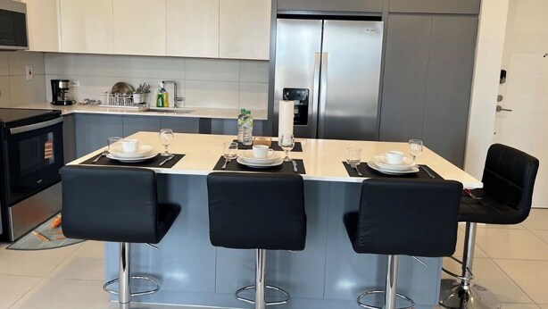 Uma cozinha moderna com uma ilha central, três banquetas de bar, uma geladeira de aço inoxidável e armários brancos.