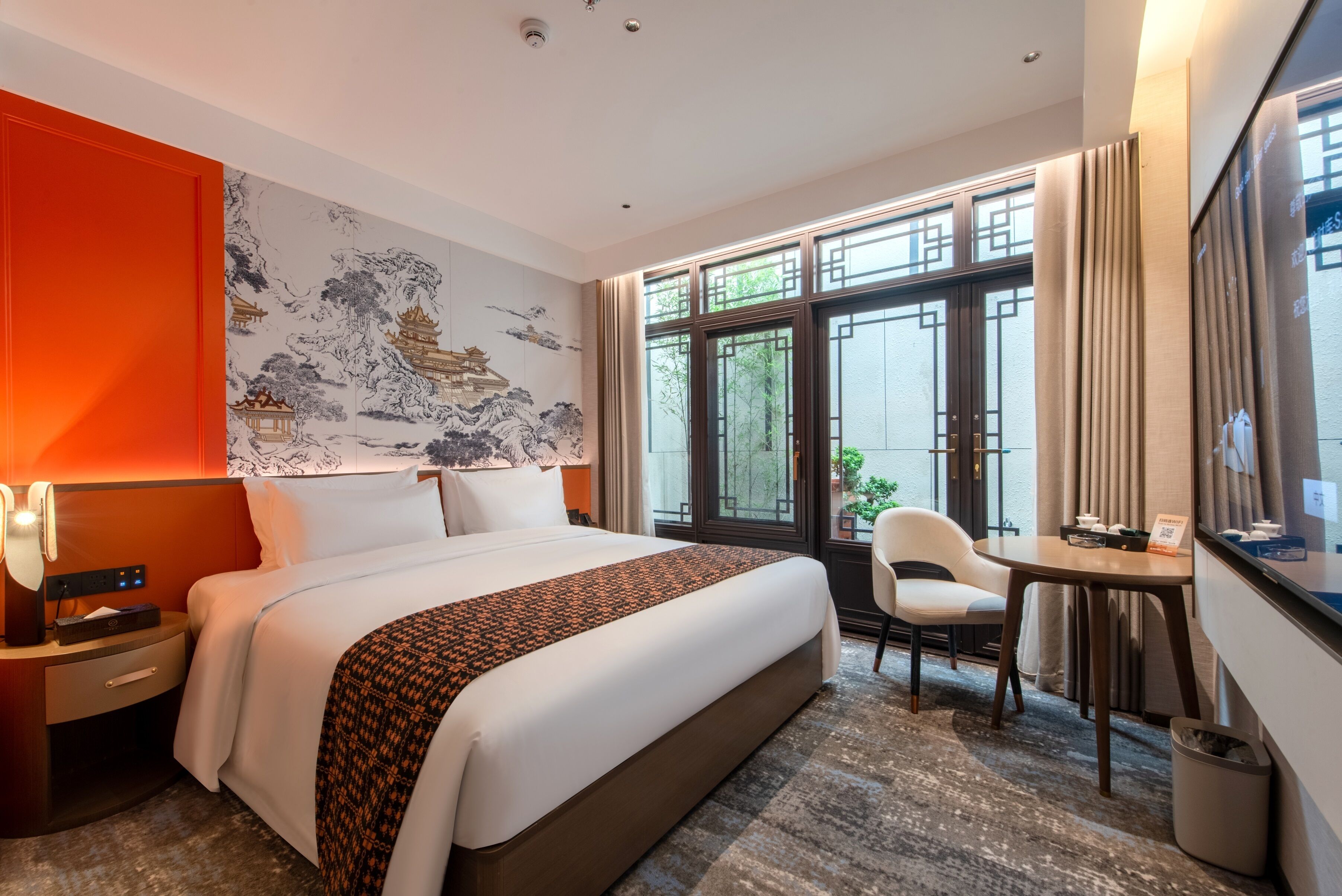 A hotel room with a large bed, a desk, a chair, and a view of greenery through the windows.