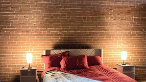 Une chambre avec un mur en briques, un lit avec une literie rouge et bleue, deux tables de chevet avec des lampes et un ventilateur de plafond.