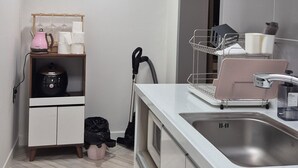 A compact kitchen with a white countertop, a built-in oven, and a stainless steel sink. There is a wooden shelf with a kettle and glassware, and a vacuum cleaner is leaning against the wall. - pacific (Gyeongju)