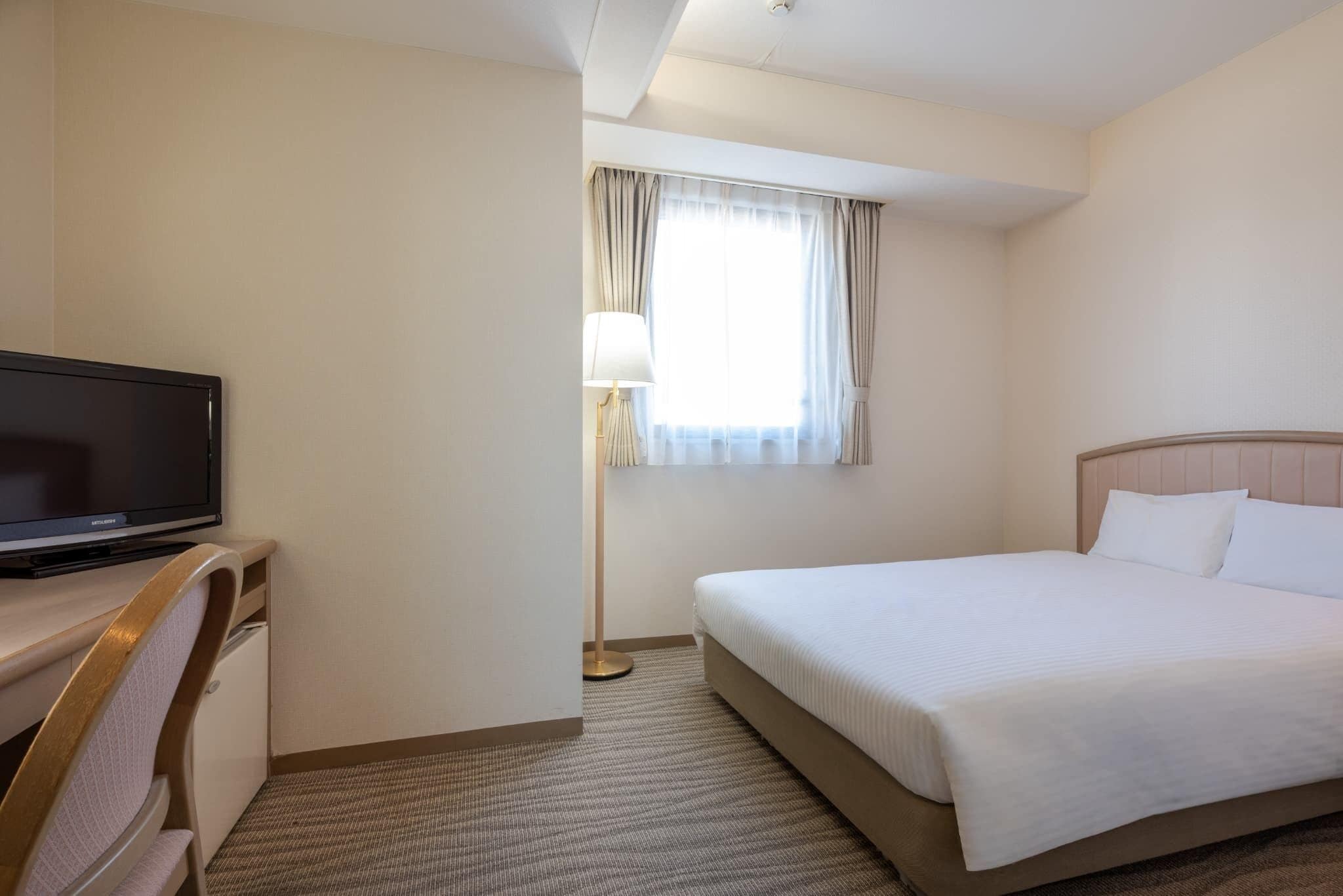 Une chambre d’hôtel avec un lit, un bureau avec une télévision, une lampe et une fenêtre avec des rideaux.