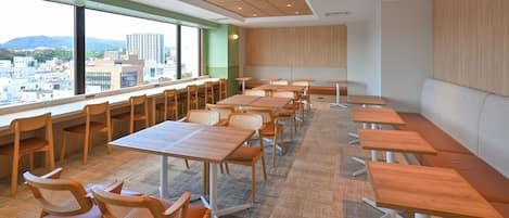 A modern conference room with wooden tables and chairs, large windows offering a city view, and a high ceiling with recessed lighting.