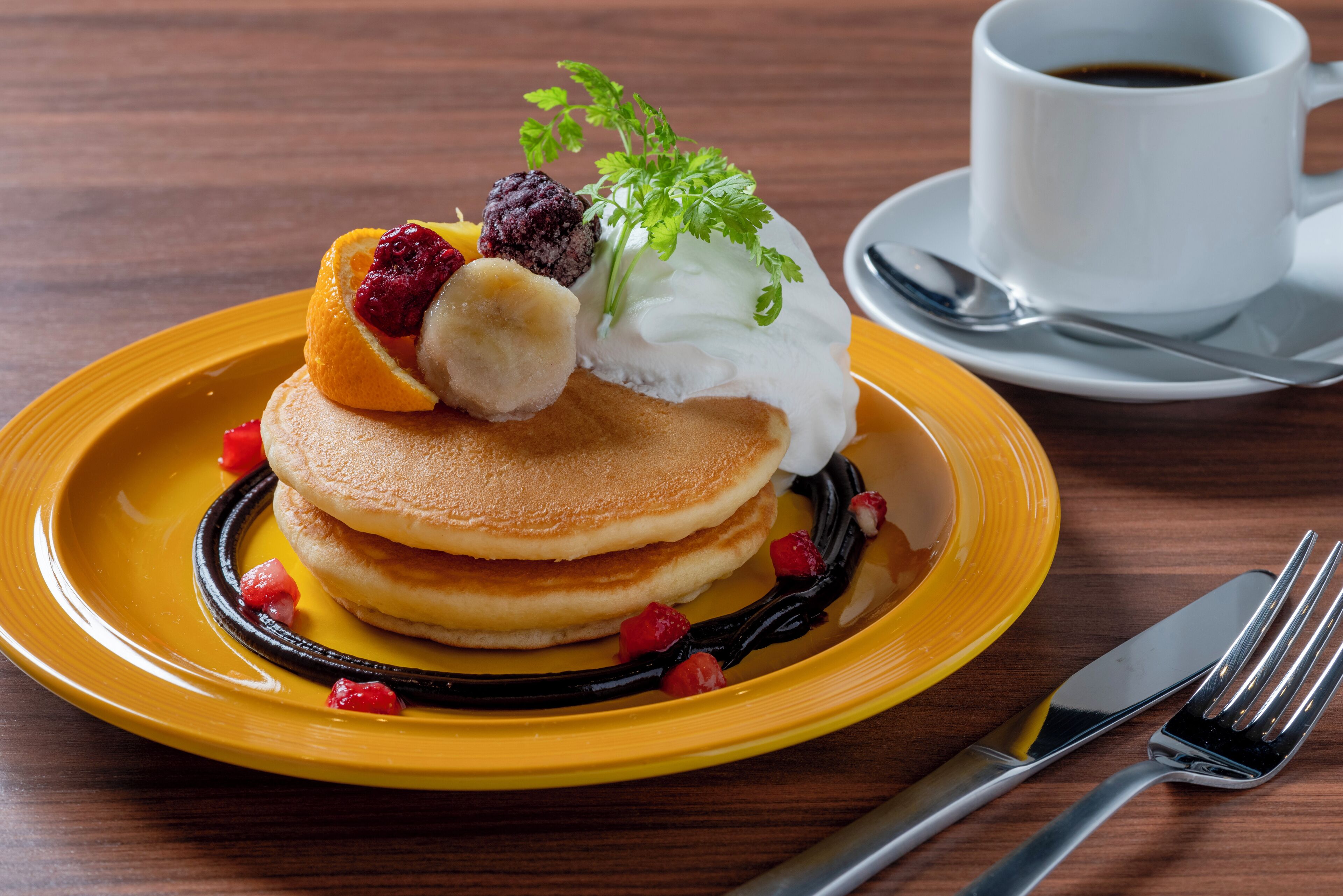Un plato de panqueques con fruta y crema, una taza de café y cubiertos sobre una mesa de madera.
