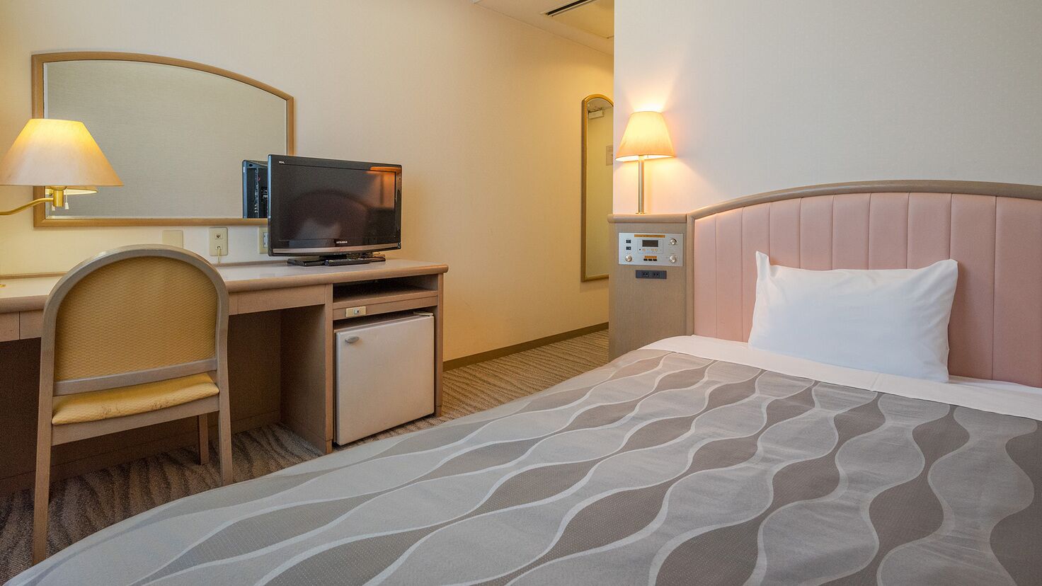 A hotel room with a bed, a desk with a television, a chair, a small refrigerator, and a bedside table with a lamp.
