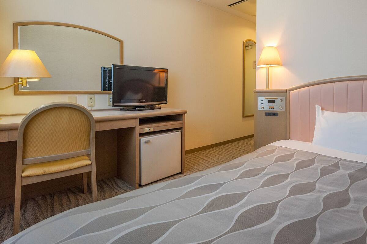 Une chambre d’hôtel comprenant un lit, un bureau avec une télévision, une chaise, un petit réfrigérateur et une table de chevet avec une lampe.