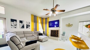 A modern living room with a sectional sofa, a dining area with yellow chairs, a fireplace, and a ceiling fan. - Modern & Cozy Retreat Game Room, Family-Friendly (North Las Vegas)