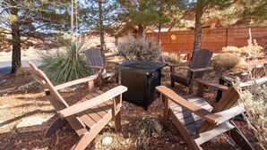Een terras met vier Adirondack-stoelen en een vierkante tafel, omgeven door woeste plantengroei en een bakstenen muur.