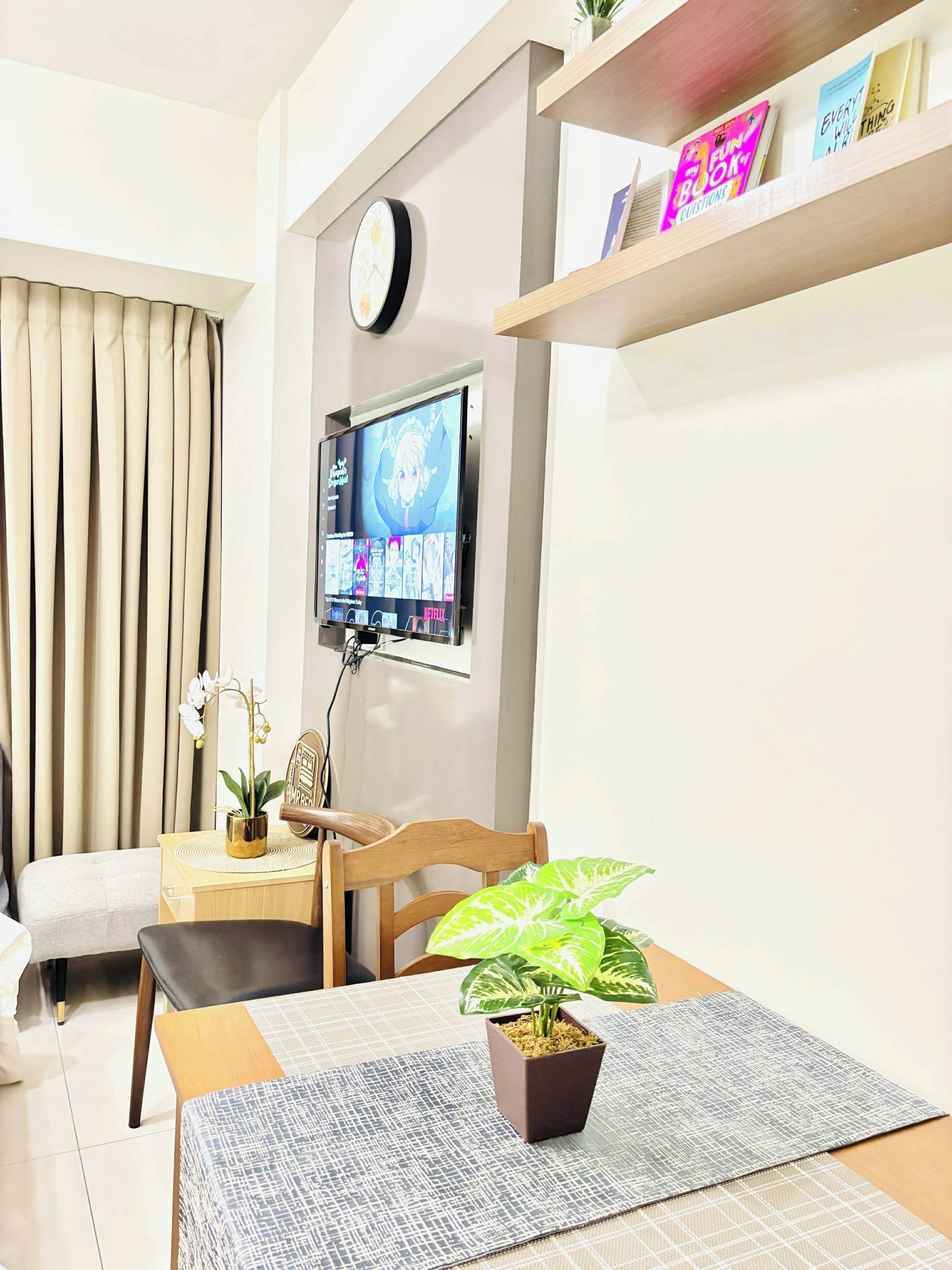 A modern living room with a flat-screen TV mounted on the wall, a dining table with a potted plant, a sofa, and a decorative clock.