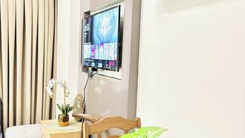 A modern living room with a flat-screen TV mounted on the wall, a dining table with a potted plant, a sofa, and a decorative clock.