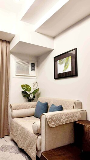 A cozy room with a sofa, a framed picture, a window with blinds, and a plant. - MR BED-Skyline View Condo With POOL (Cebu City)