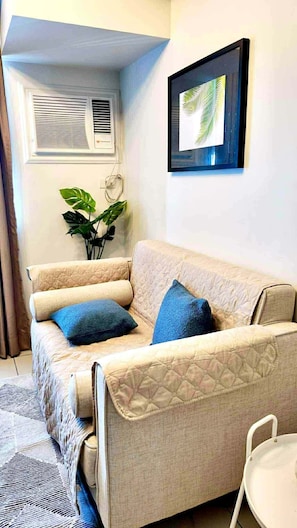 Una sala de estar con un sofá beige, dos cojines azules, un cuadro enmarcado, una planta en maceta y una silla blanca.