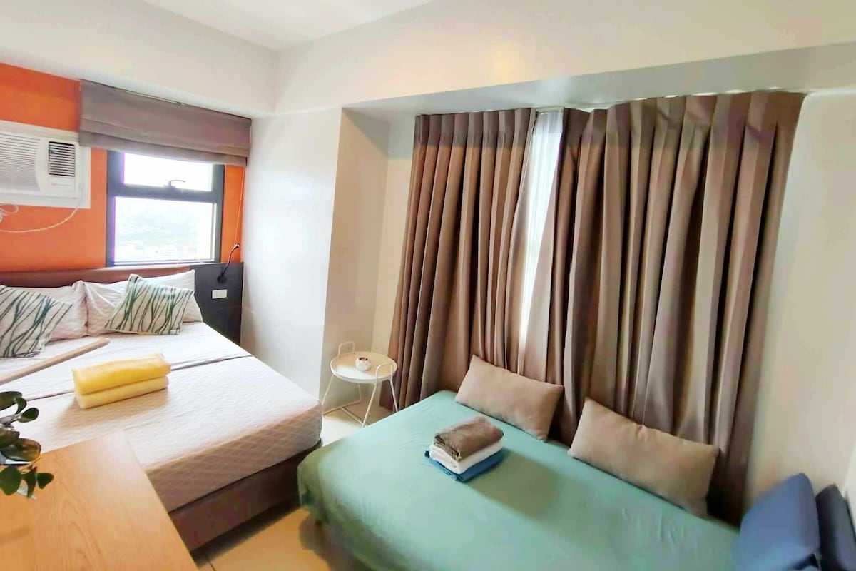 A hotel room with a bed, a bedside table, a small table with a chair, and a window with curtains.