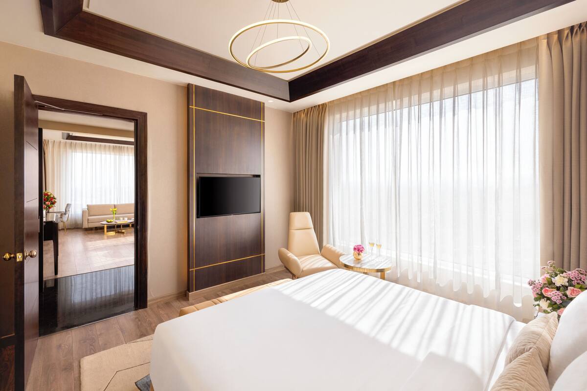 A hotel room with a large bed, a TV mounted on the wall, a chair, a small table with a vase, and a large window with curtains.