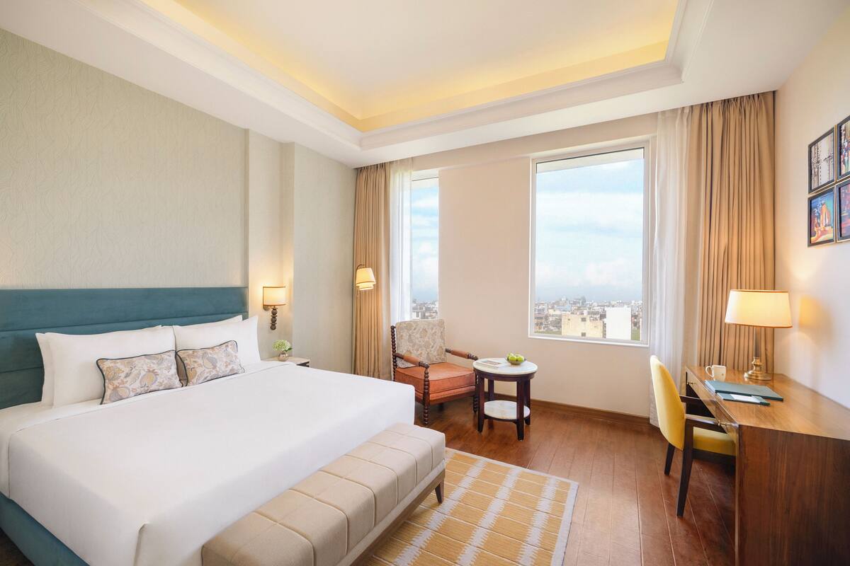A hotel room with a large bed, a desk, a chair, a small table, and a view of the city.
