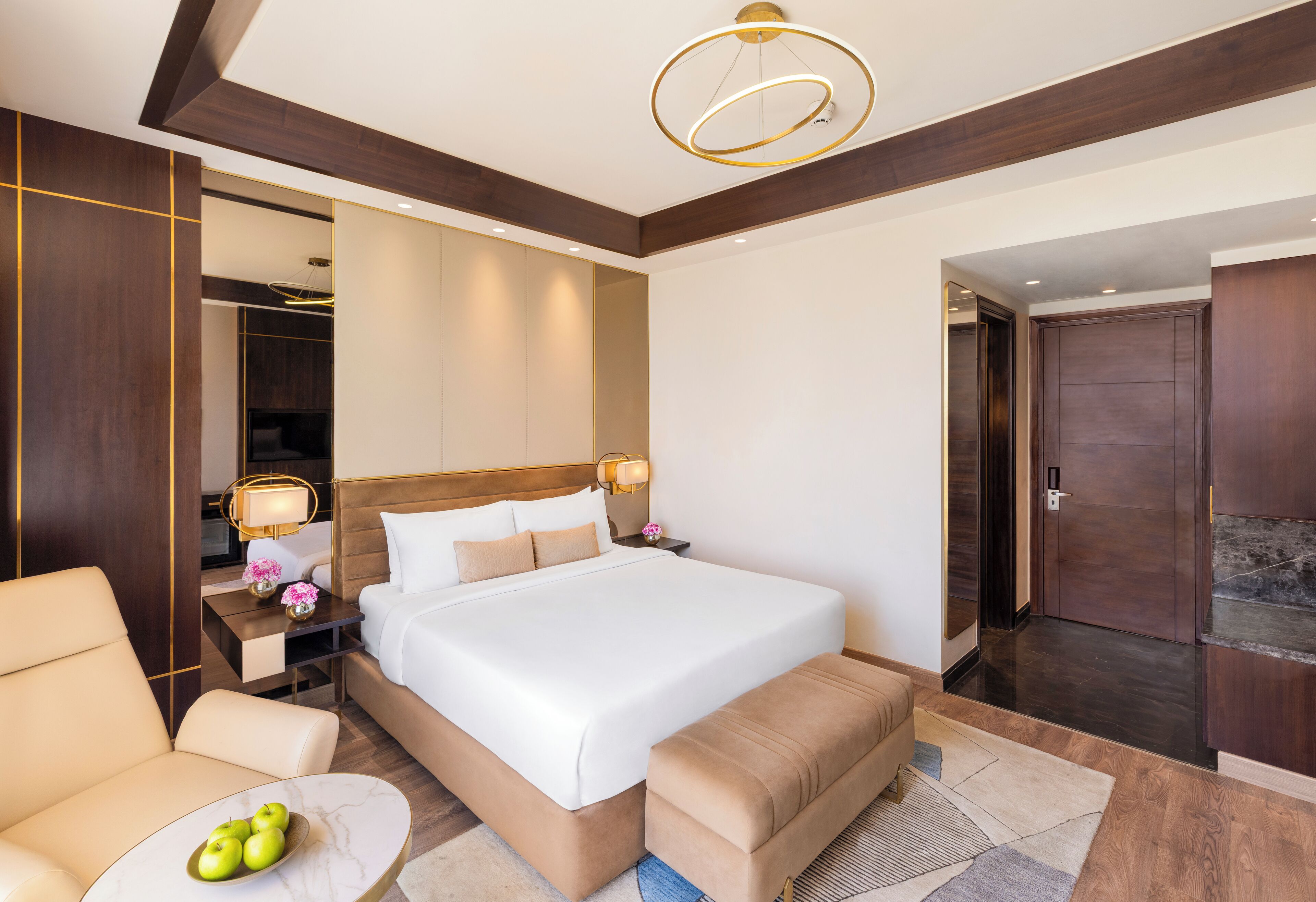 Una habitación de hotel con una cama grande, mesitas de noche, una silla, una pequeña mesa con manzanas y un espejo.
