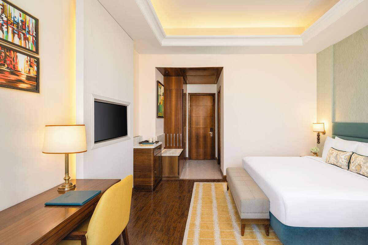 A hotel room with a bed, desk, chair, TV, artwork, and a door leading to a bathroom.