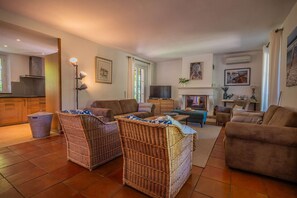 Ein Wohnzimmer mit Korbmöbeln, einem Kamin, einem Fernseher und einer durchgehenden Tür erkennbaren Küchenzeile.