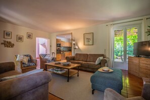 Ein Wohnzimmer mit einer Couch, Sesseln, einem Couchtisch und einem Fernseher. Im Hintergrund ist eine Küche zu sehen.