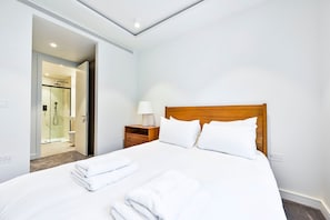 A hotel room with a bed, a nightstand, a lamp, a wooden headboard, and a shower area. - Guestready - Stylish Living in Paddington (London)