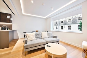 A modern living room with a grey sofa, a round coffee table, and a large window with a city view. - Guestready - Stylish Living in Paddington (London)