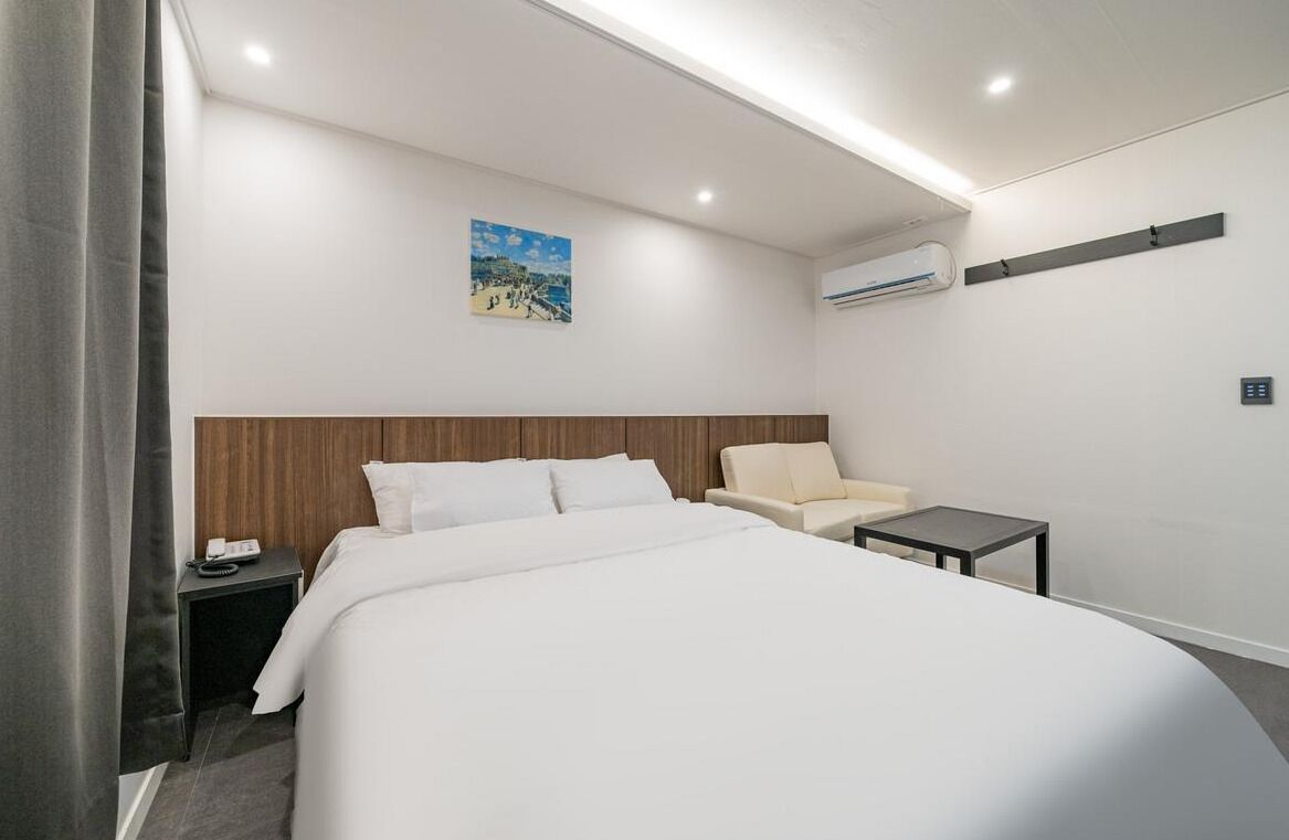 A hotel room with a bed, a chair, a small table, an air conditioning unit, and a painting on the wall.