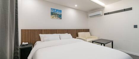A hotel room with a bed, a chair, a small table, an air conditioning unit, and a painting on the wall.