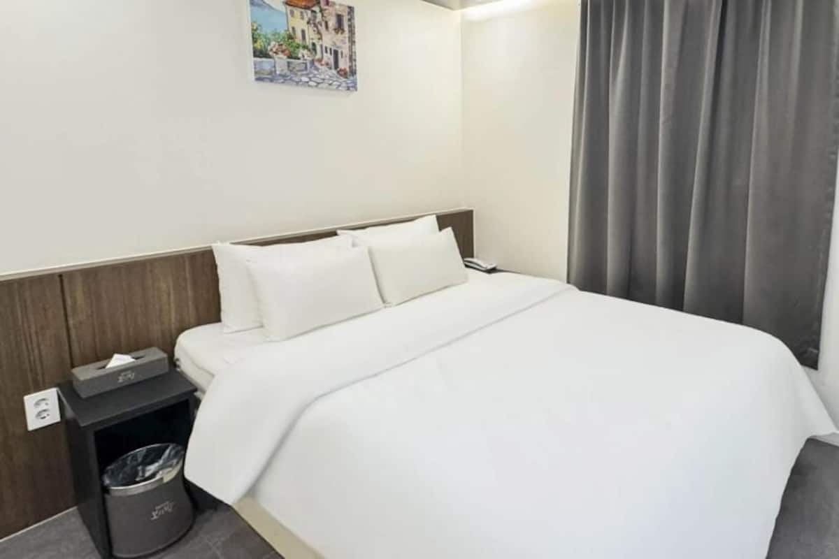 Una habitación de hotel con una cama, una mesita de noche, un cesto de basura, un escritorio y una cortina.