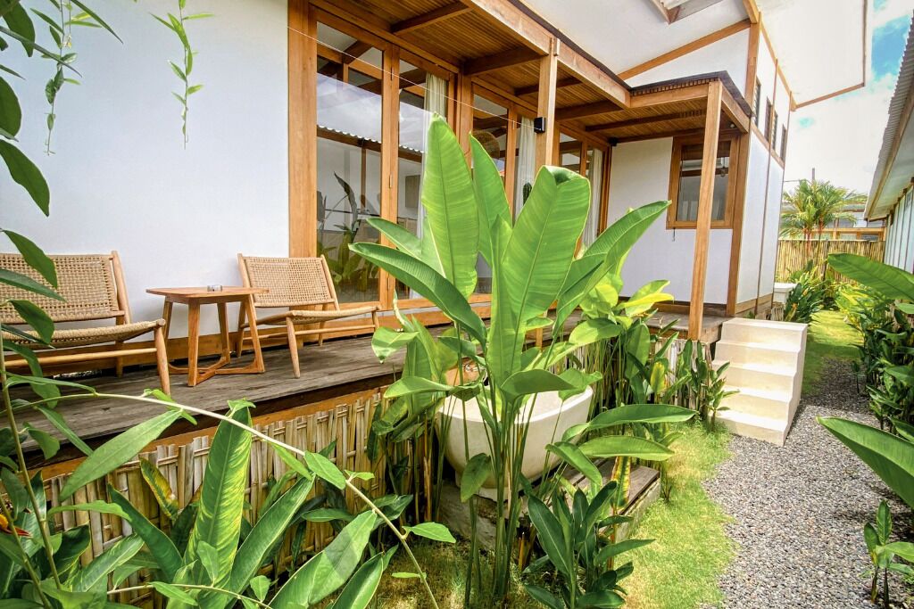 A wooden deck with two wicker chairs and a table, surrounded by lush greenery and a white wall with windows.