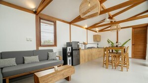 Un salon moderne comprenant un canapé, une table basse en bois, une table à manger avec des chaises, un coin cuisine équipé d’appareils électroménagers et une grande fenêtre avec des stores.