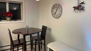 Een eethoek met een ronde tafel, vier stoelen, een wandklok, een windmolen decoratie en een raam met rode bloemen.