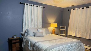 Een slaapkamer met een bed, nachtkastje, lamp en gordijnen.