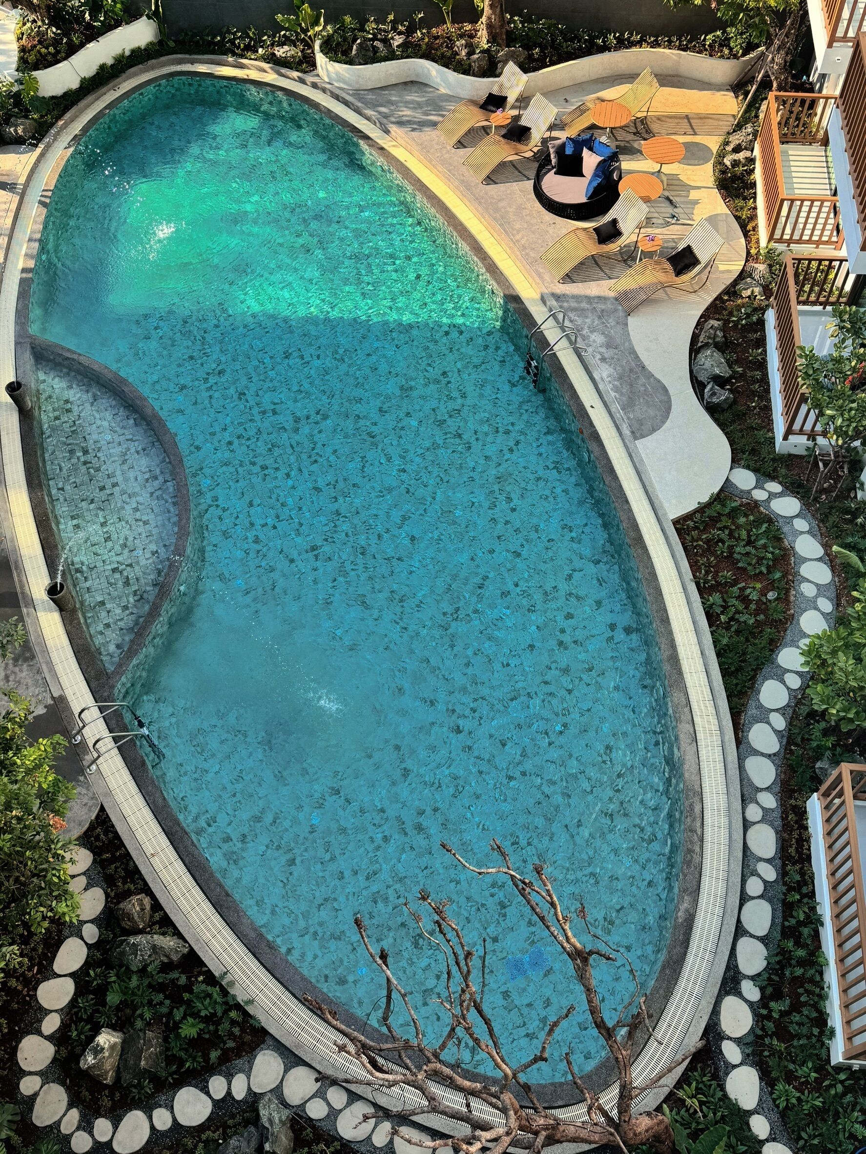An oval-shaped swimming pool with a tiled deck, lounge chairs, and a small table.
