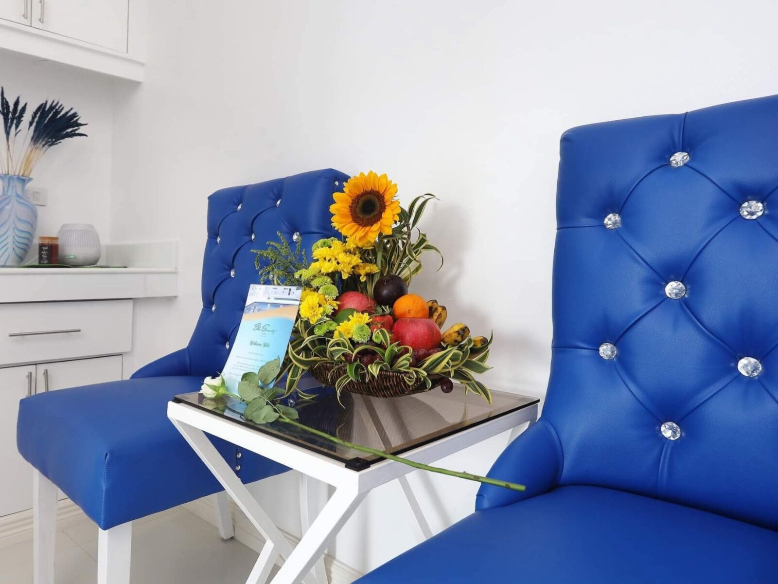 Una silla tapizada de azul con una mesita de vidrio, un florero con flores y una cesta de frutas.